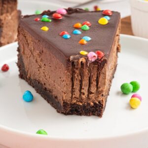 A slice of Cosmic Brownie Cheesecake topped with colorful candy sits on a white plate.
