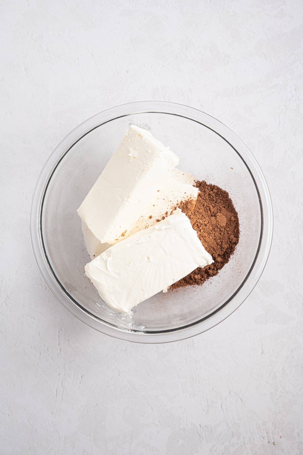 Two blocks of cream cheese and cocoa powder in a clear glass mixing bowl on a light surface.