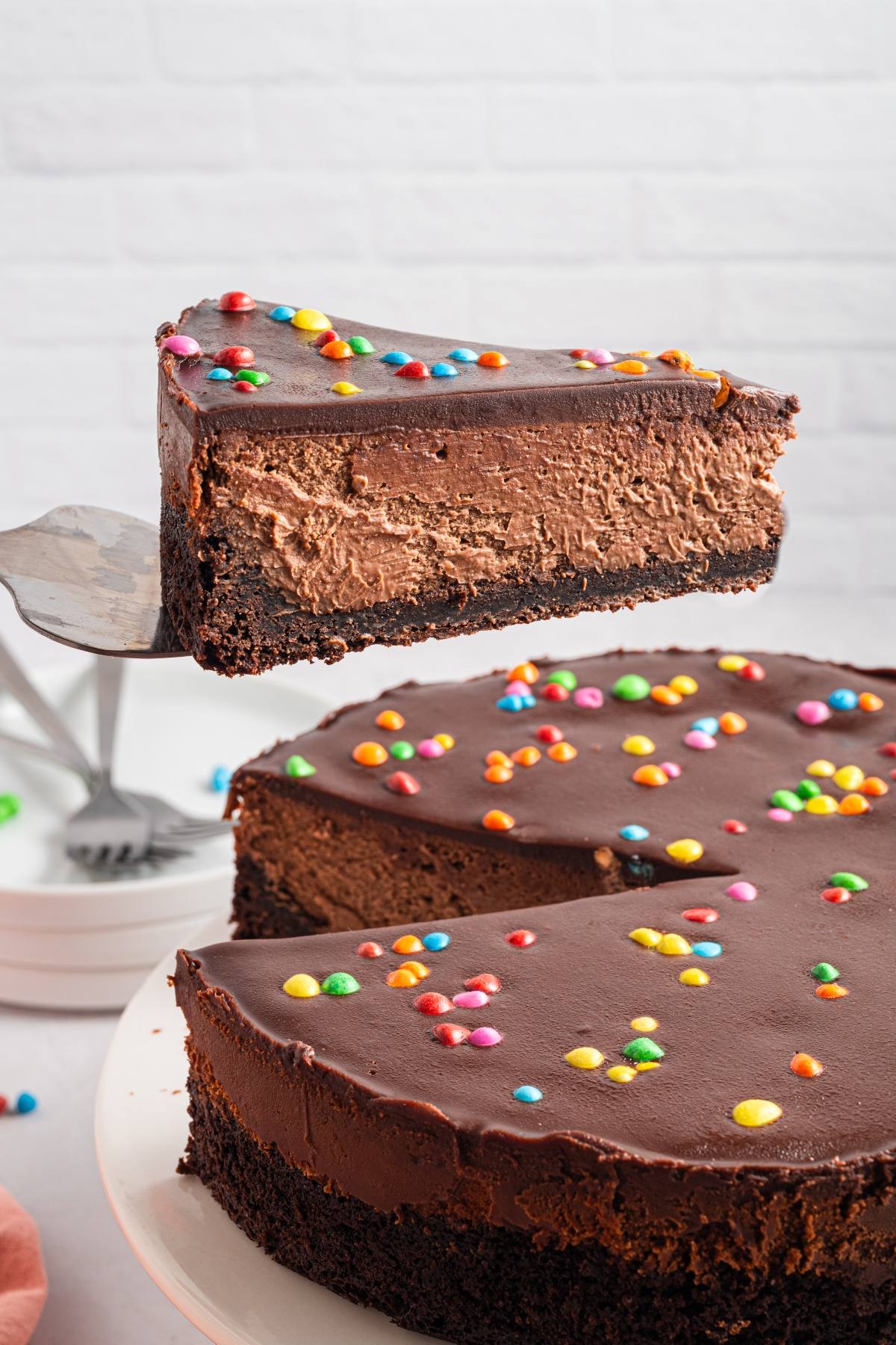 A slice of cosmic brownie cheesecake with colorful candy sprinkles being lifted from the whole cake.