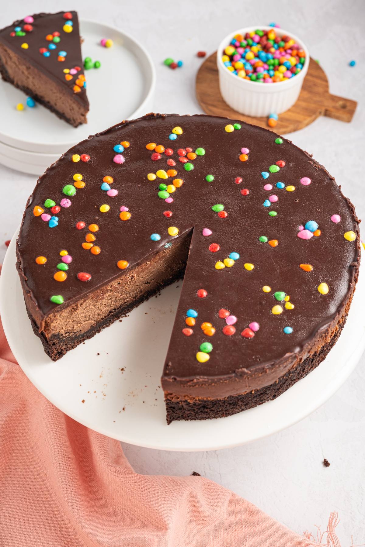 A cosmic brownie cheesecake with colorful candy sprinkles and one slice missing, on a white cake stand.