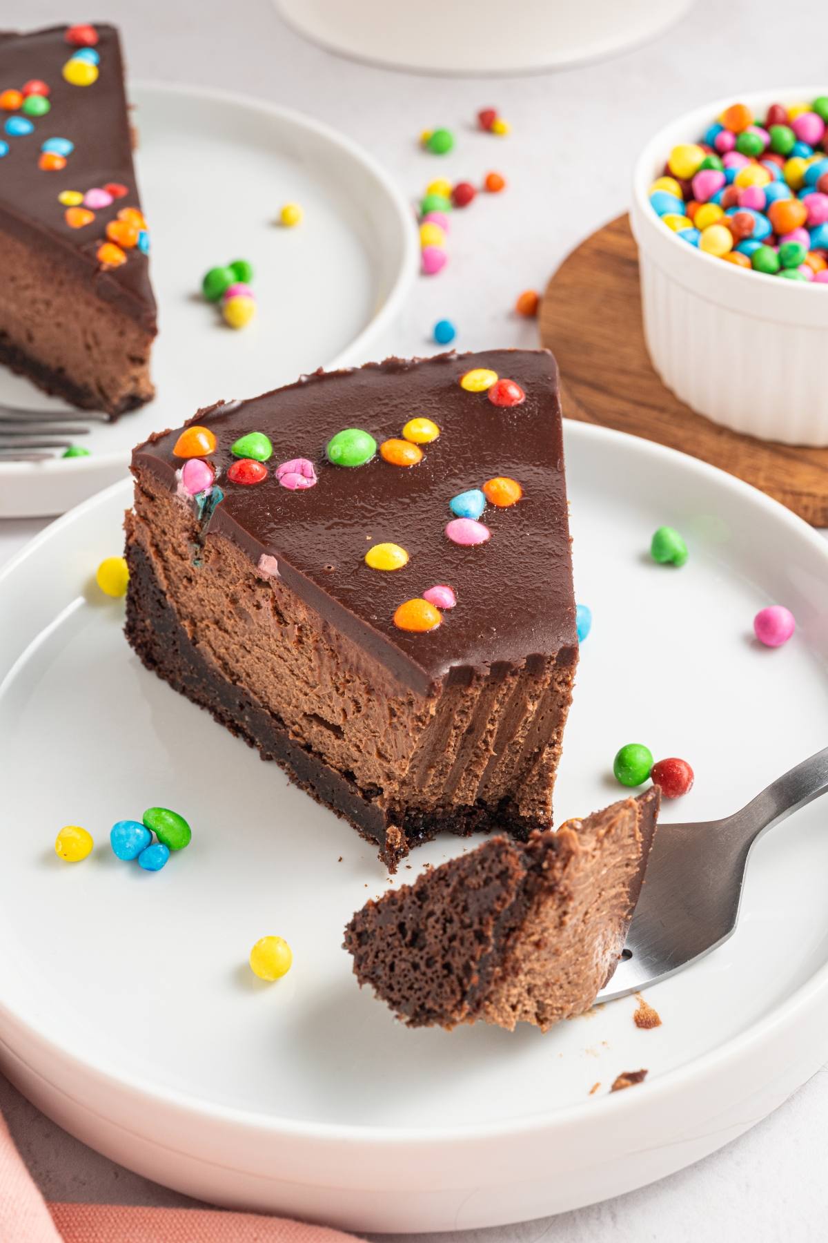 A slice of cosmic brownie cheesecake with colorful candy on top, served on a white plate with a forkful cut out.