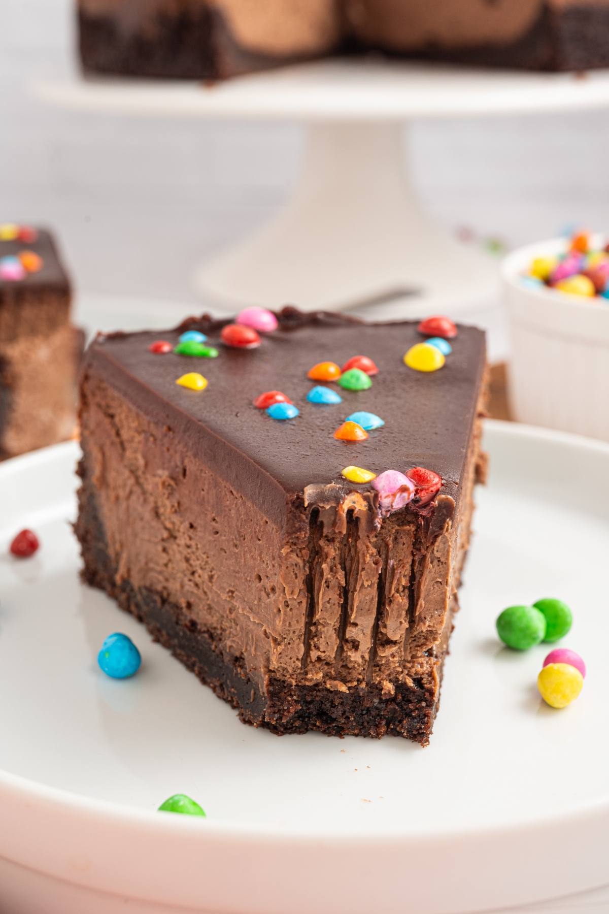 A slice of cosmic brownie cheesecake with colorful candy toppings on a white plate.