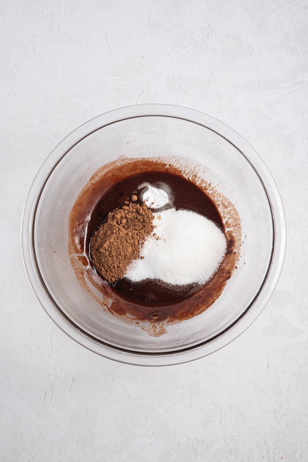 A glass bowl with cocoa powder, sugar, and liquid chocolate mixture on a white surface.