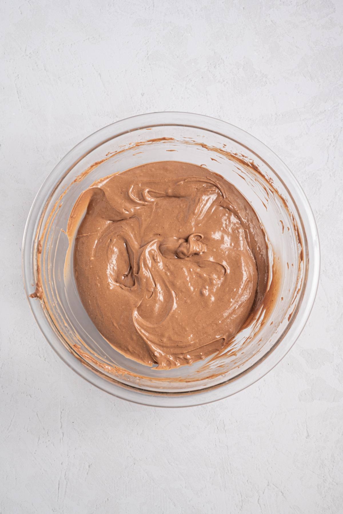 A glass bowl filled with smooth chocolate cake batter on a light textured surface.