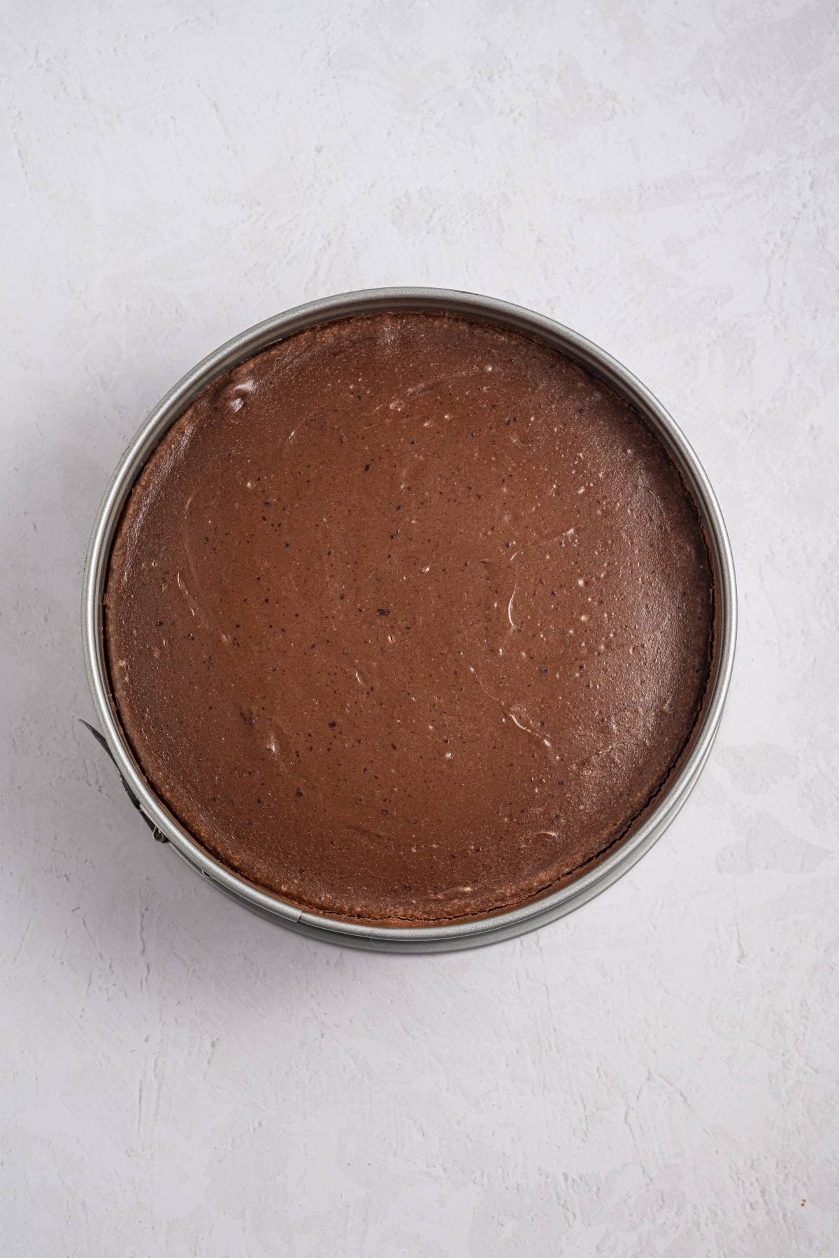 Round chocolate cake in a metal pan on a light textured surface, viewed from above.