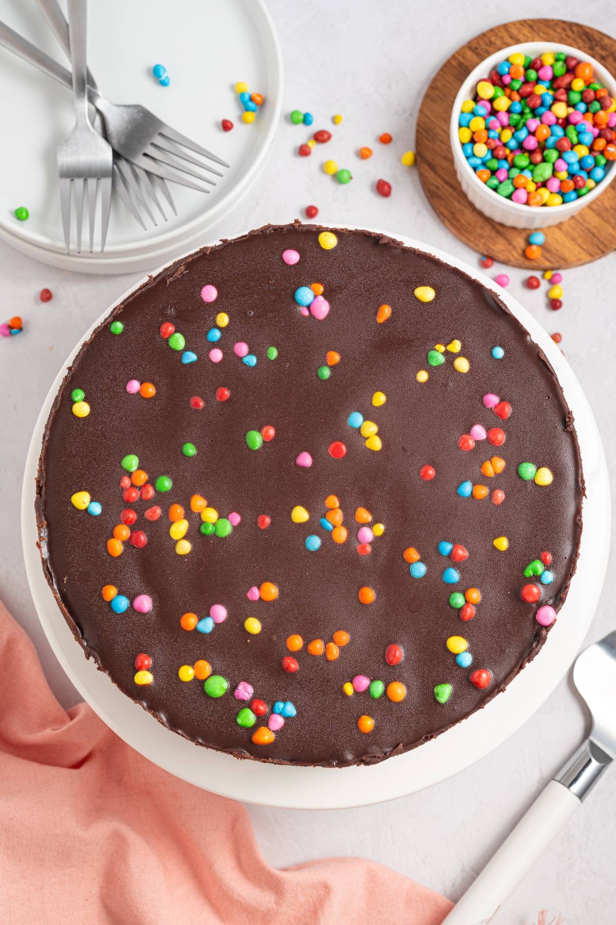A cosmic brownie cheesecake with colorful candy sprinkles, next to forks, a bowl of sprinkles, and a pink napkin.