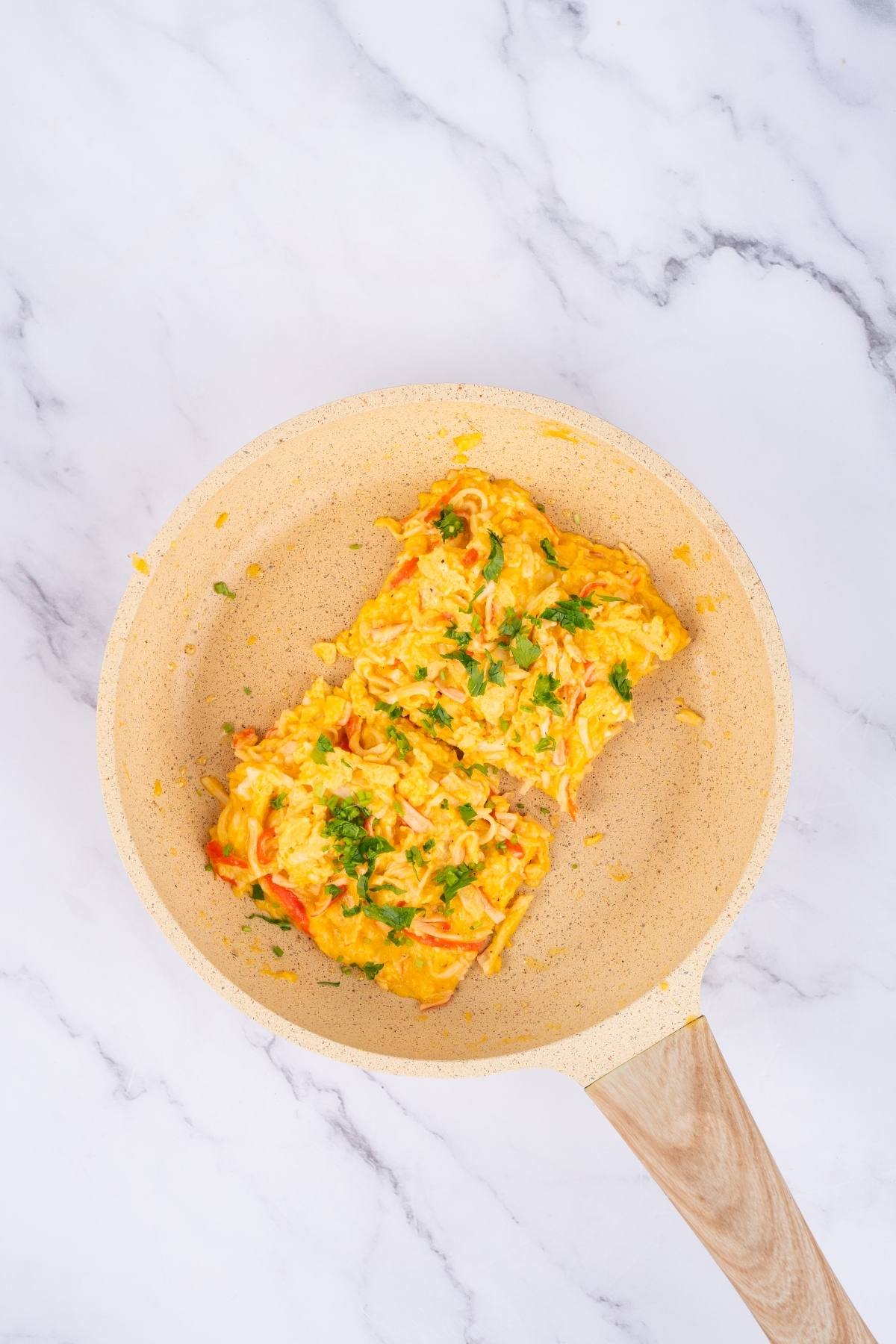 Scrambled eggs with herbs in a light-colored frying pan on a marble surface.