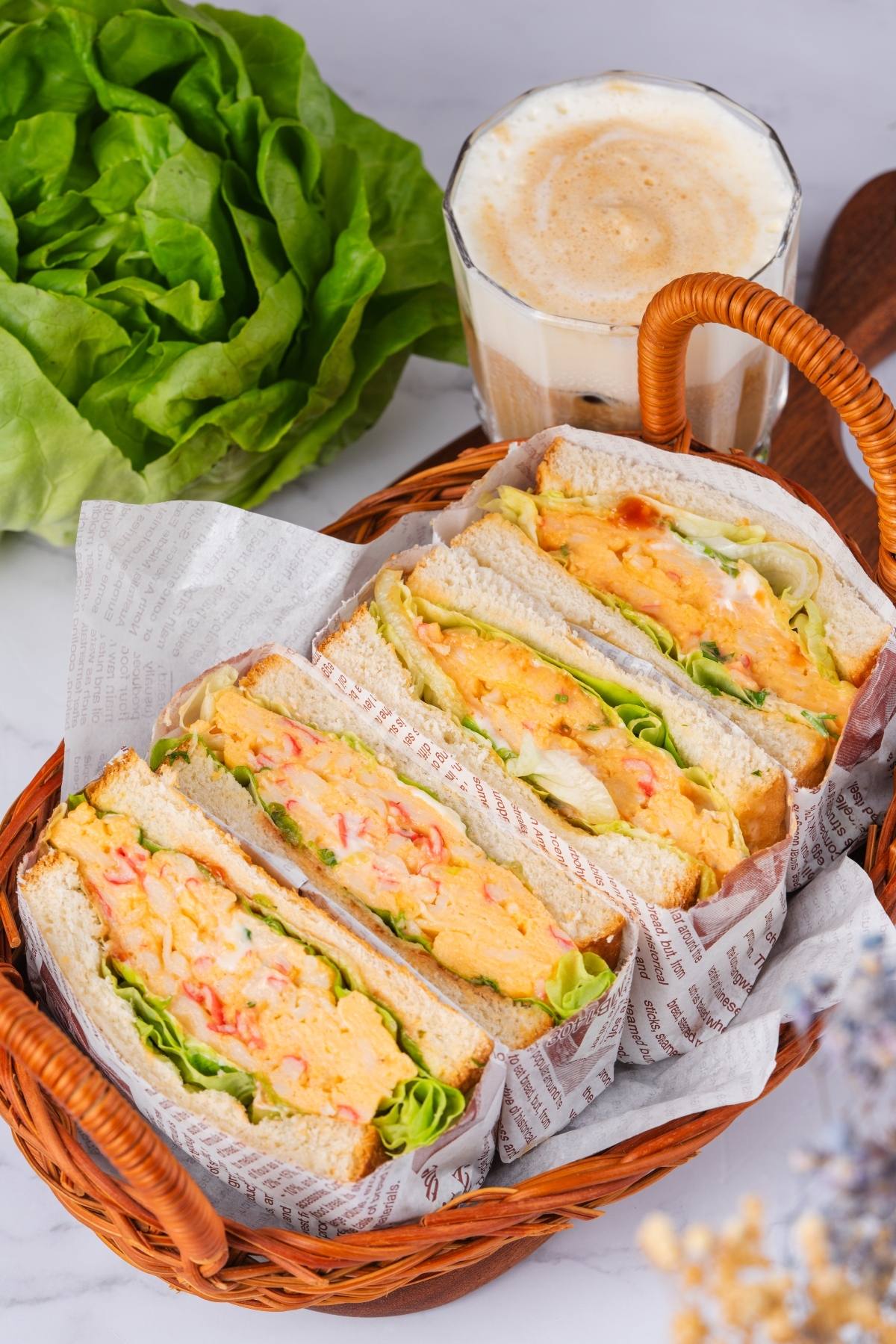 Basket with egg salad sandwiches, lettuce, and a glass of coffee on a marble surface.