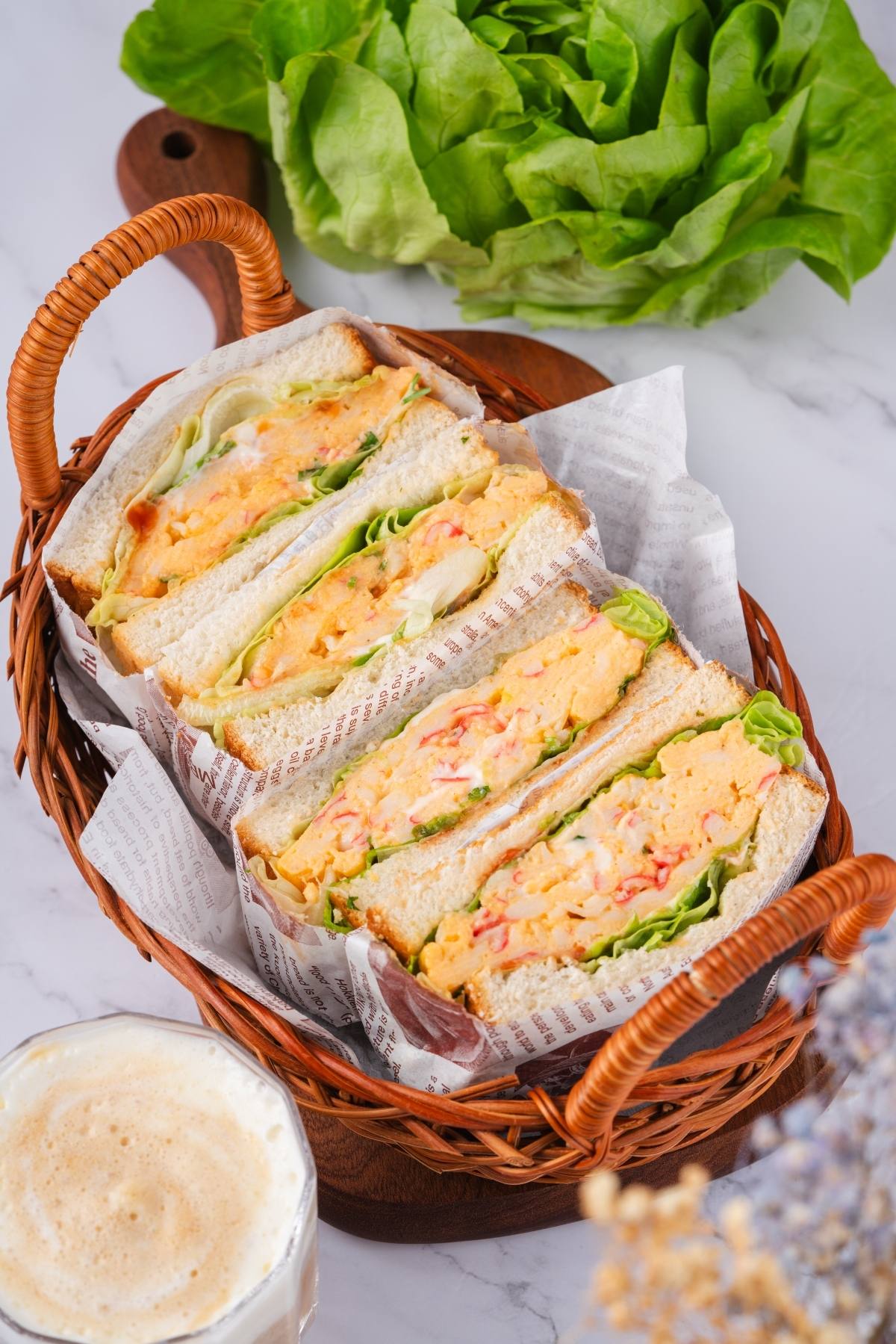 Three sandwiches with lettuce and fillings in a wicker basket, next to a leafy lettuce and a drink.