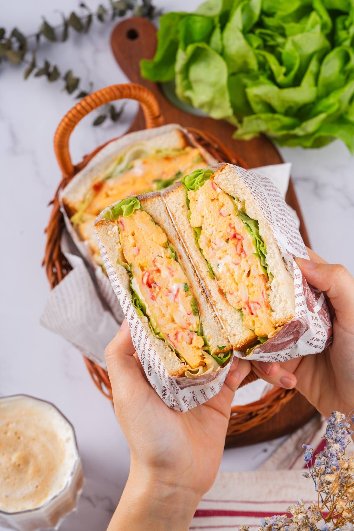 Hands holding a sandwich with egg salad, lettuce, and vegetables; basket and greens in background.