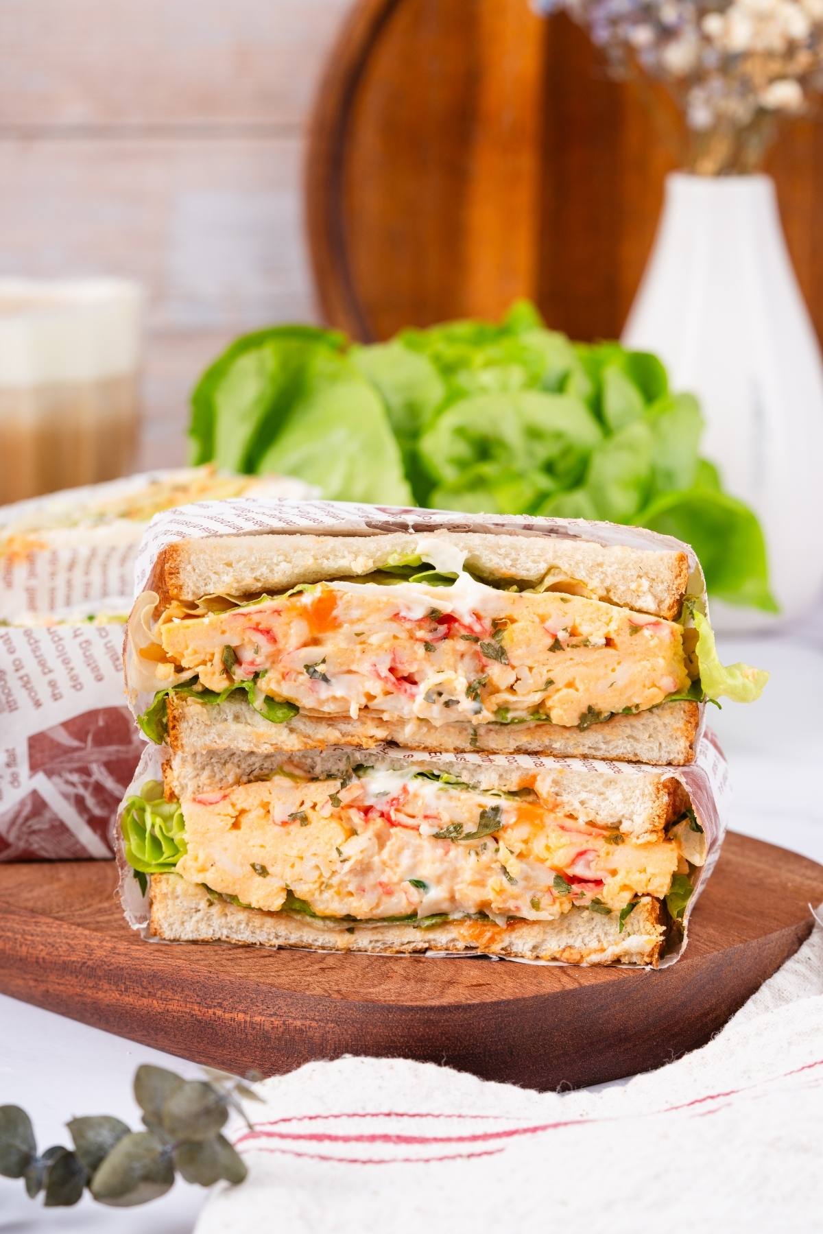 A stacked sandwich with creamy filling, lettuce, and bread, displayed on a wooden board with greens behind.