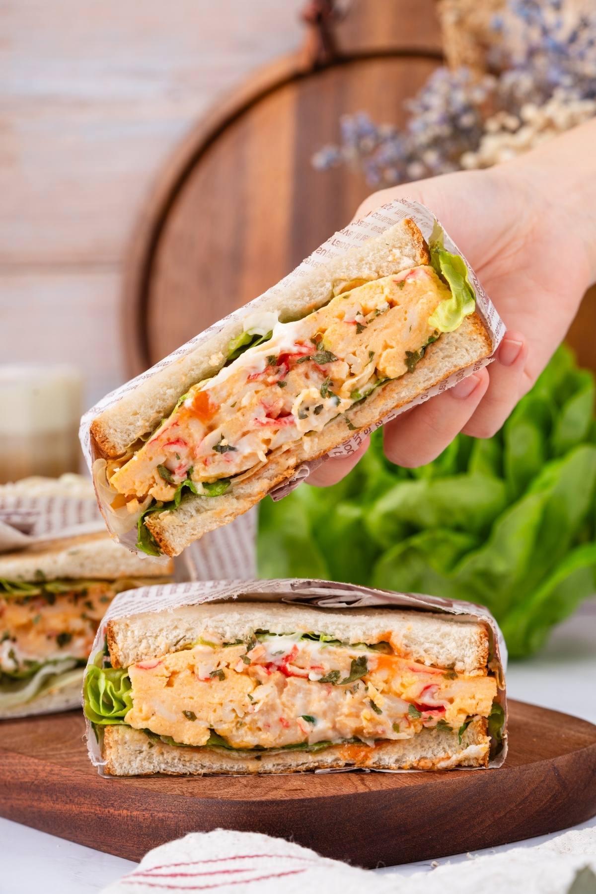 Hand holding a thick, vegetable-filled sandwich with lettuce and creamy spread, with another on a wooden plate.