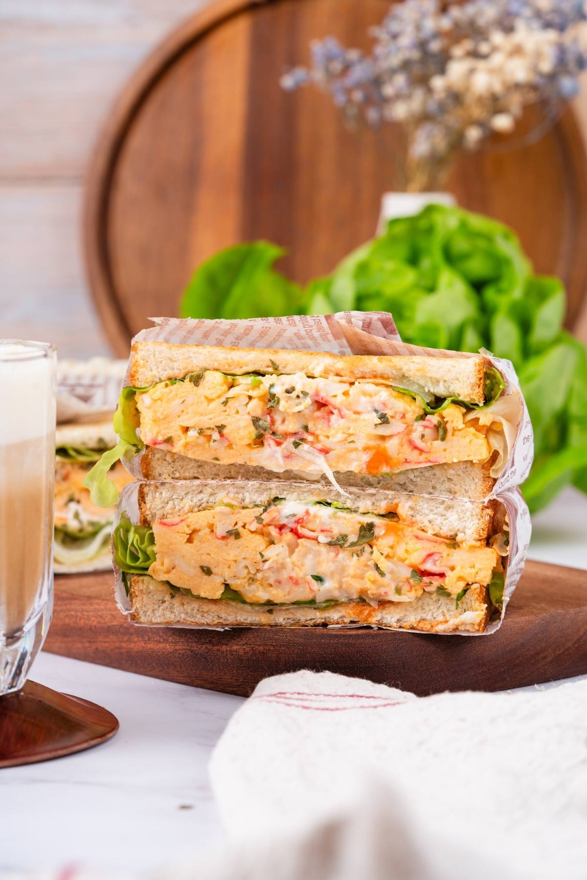A thick crab sandwich with creamy filling and lettuce, cut in half, next to a glass of coffee on a table.