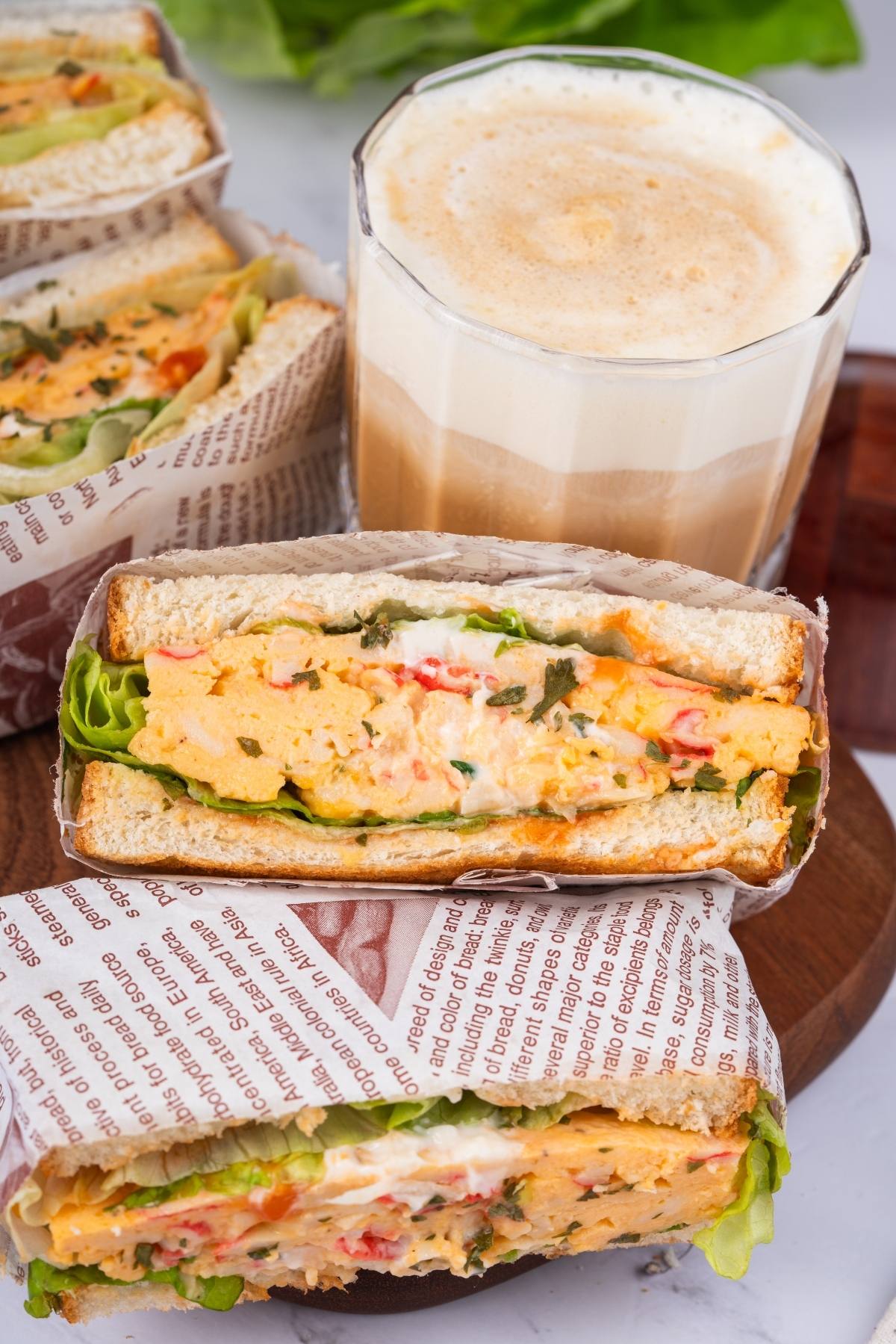 Egg salad sandwiches with lettuce, wrapped in printed paper, served with a glass of frothy coffee.