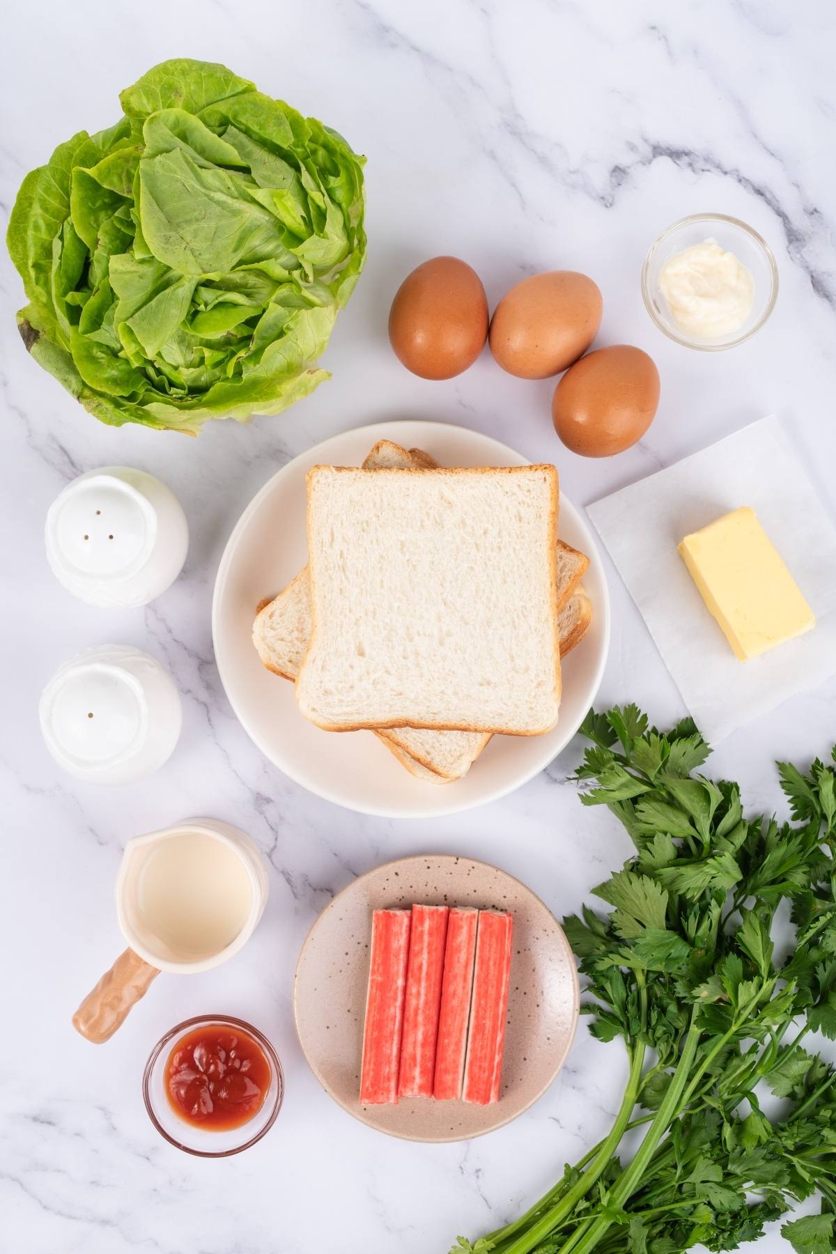 Top-down view of sandwich ingredients: bread, lettuce, eggs, butter, mayo, parsley, surimi, ketchup, milk, salt, pepper.