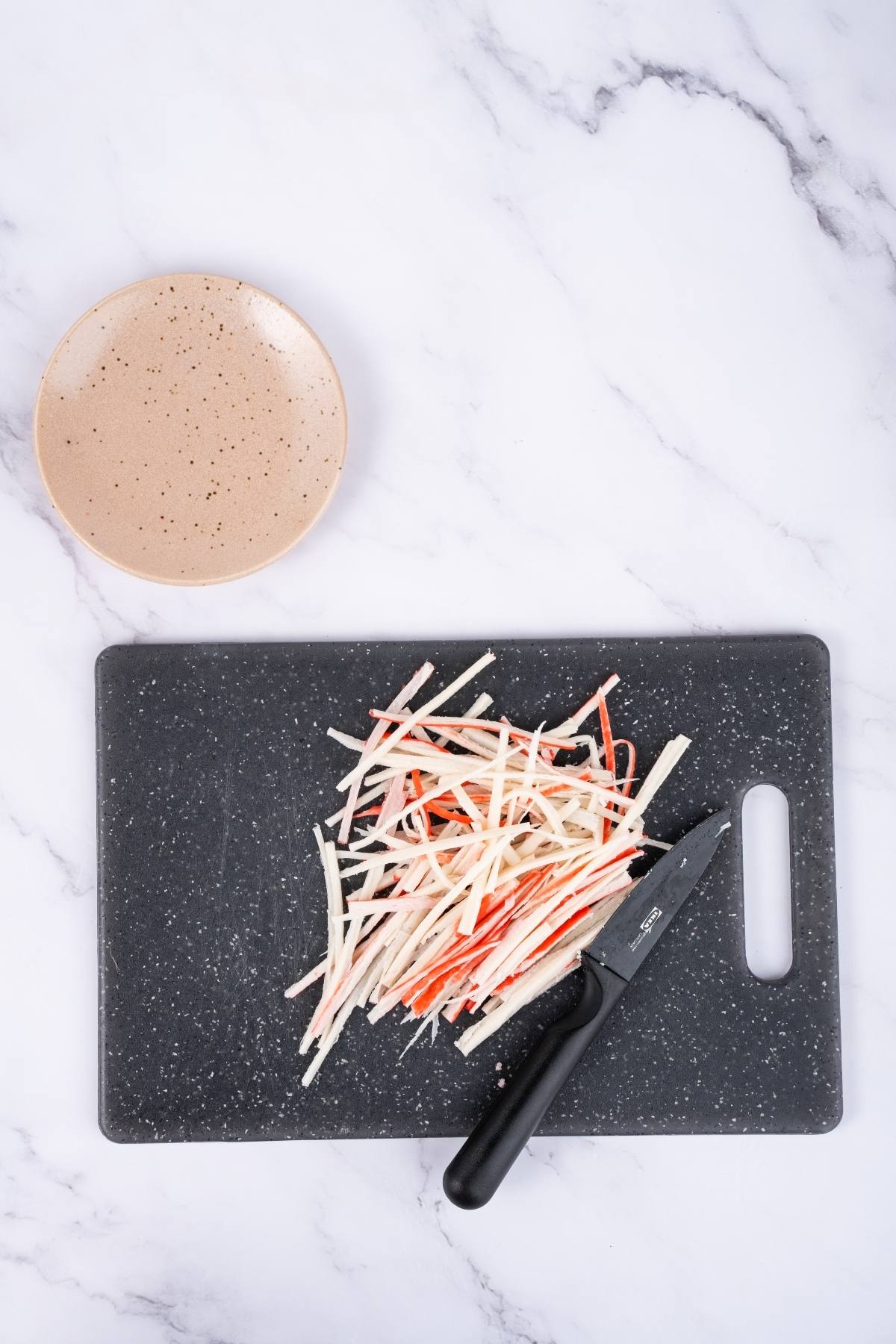 A cutting board with sliced crab sticks and a knife, next to an empty beige plate on a marble countertop.