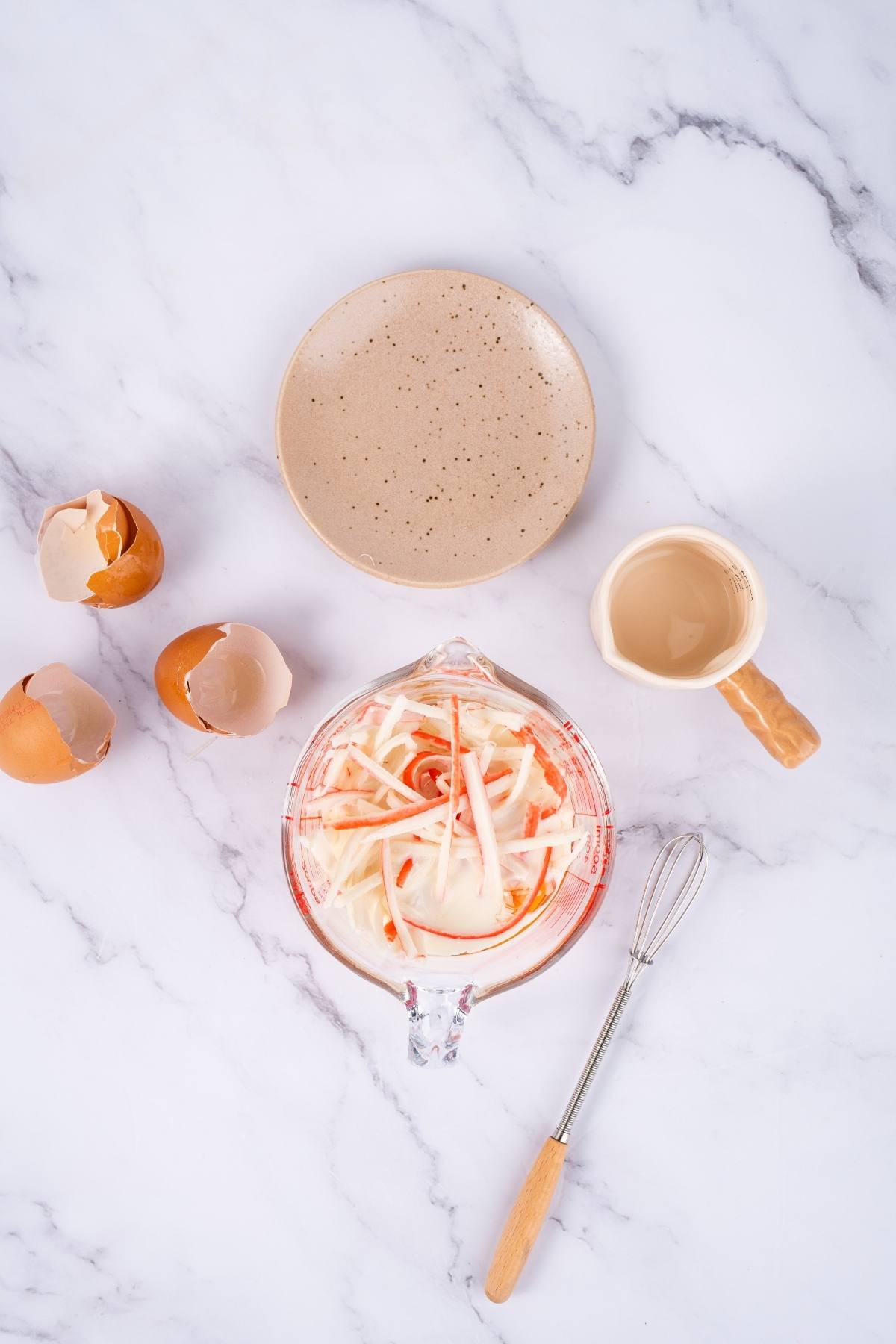 Measuring cup with sliced vegetables, empty plate, eggshells, small pitcher, and whisk on a marble surface.