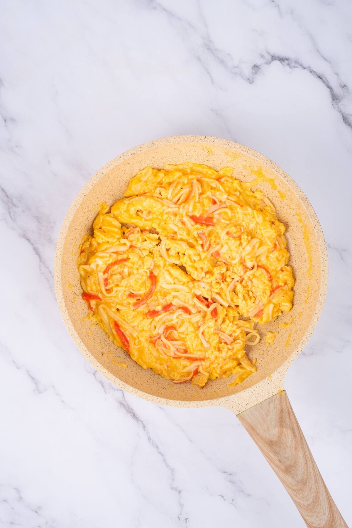 Scrambled eggs with red bell pepper in a light-colored frying pan on a marble surface.
