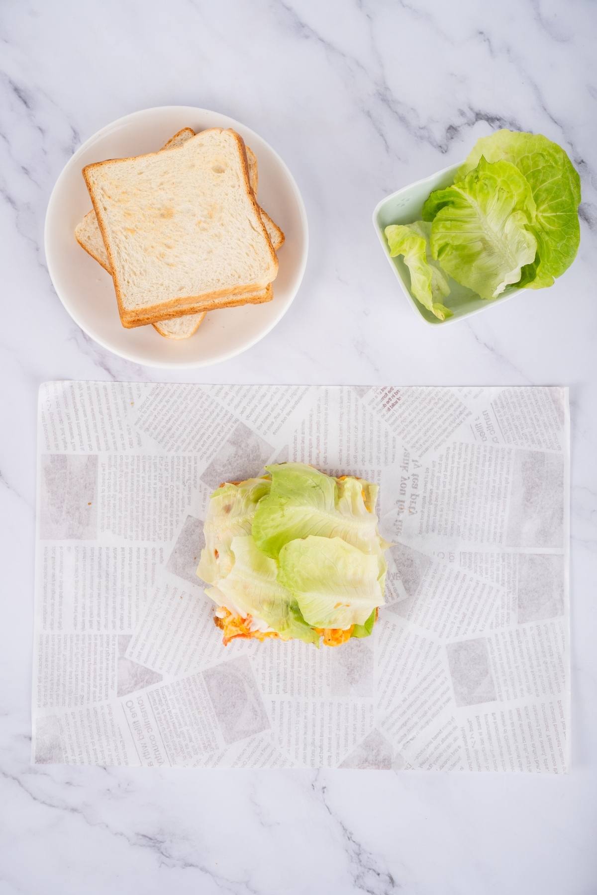 Sandwich filling with lettuce on paper, slices of bread on a plate, and lettuce in a bowl on a marble surface.