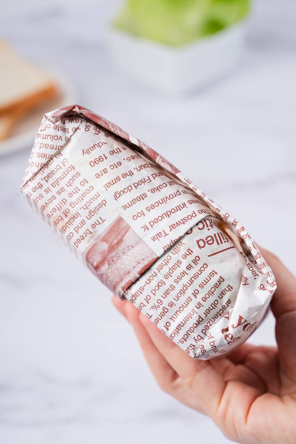 A hand holding a sandwich wrapped in printed newspaper-style paper, with blurred food in the background.