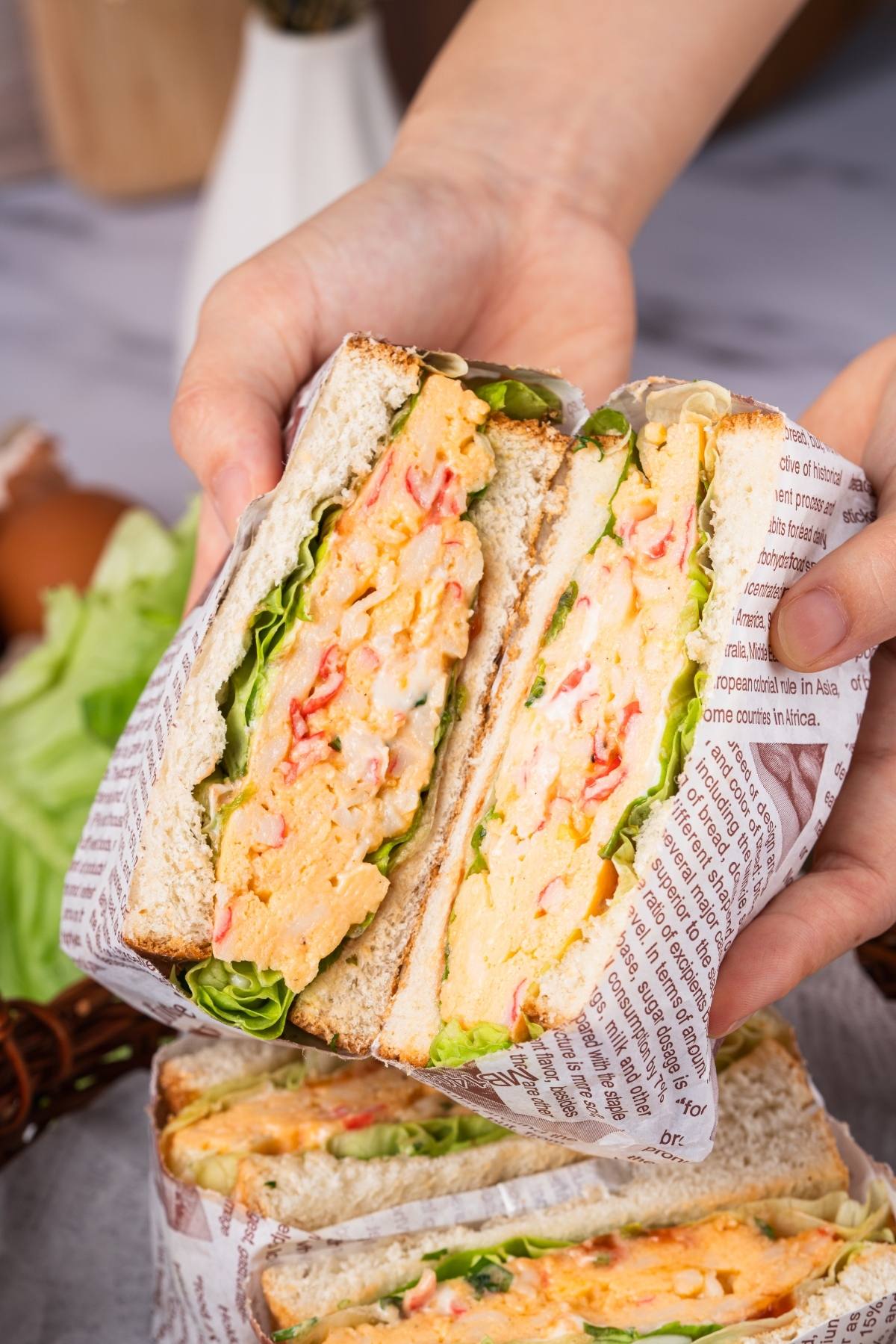 A hand holds a sandwich with egg, lettuce, and tomato, wrapped in printed paper.
