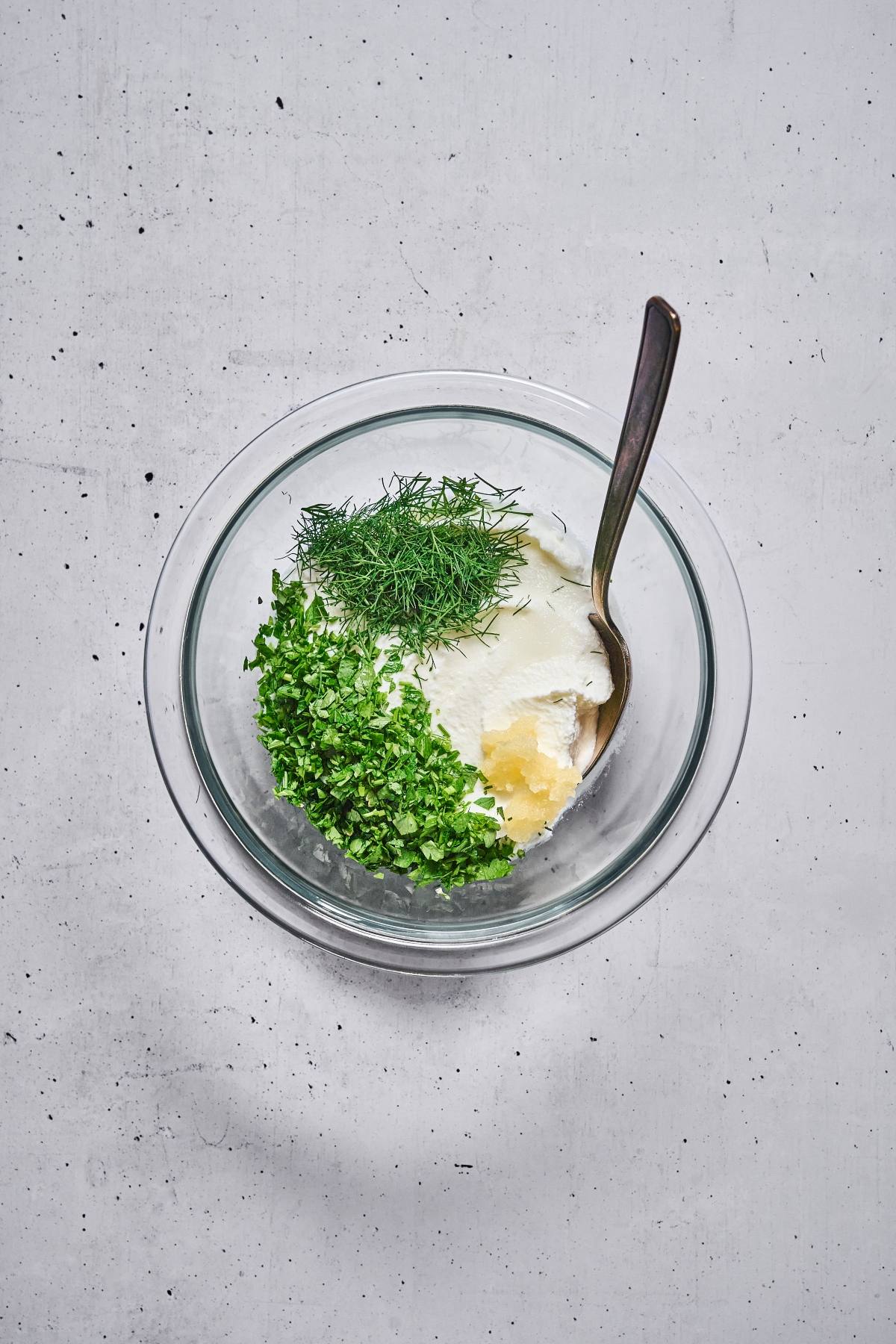 A glass bowl with sour cream, chopped parsley, dill, and a spoon on a white surface.