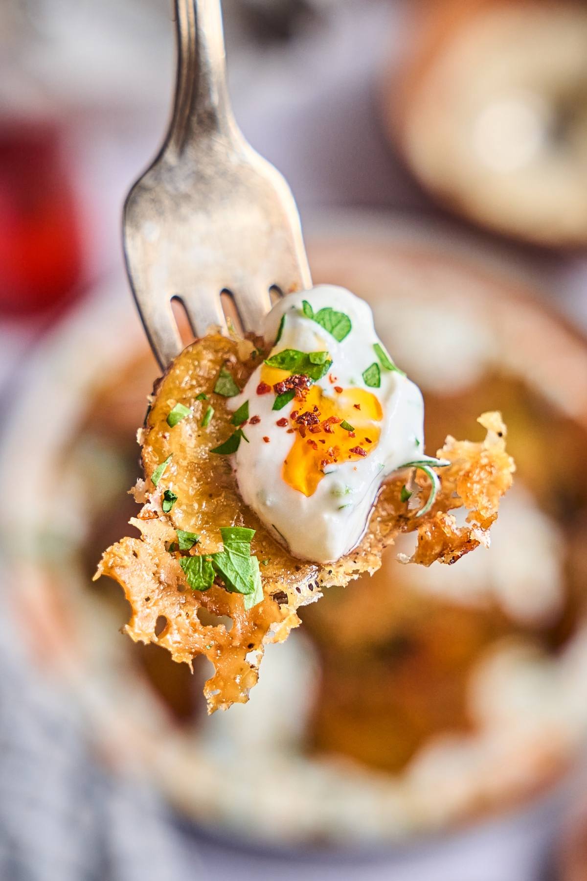 A fork holds a crispy potato slice topped with sour cream, herbs, and a sprinkle of red pepper flakes.
