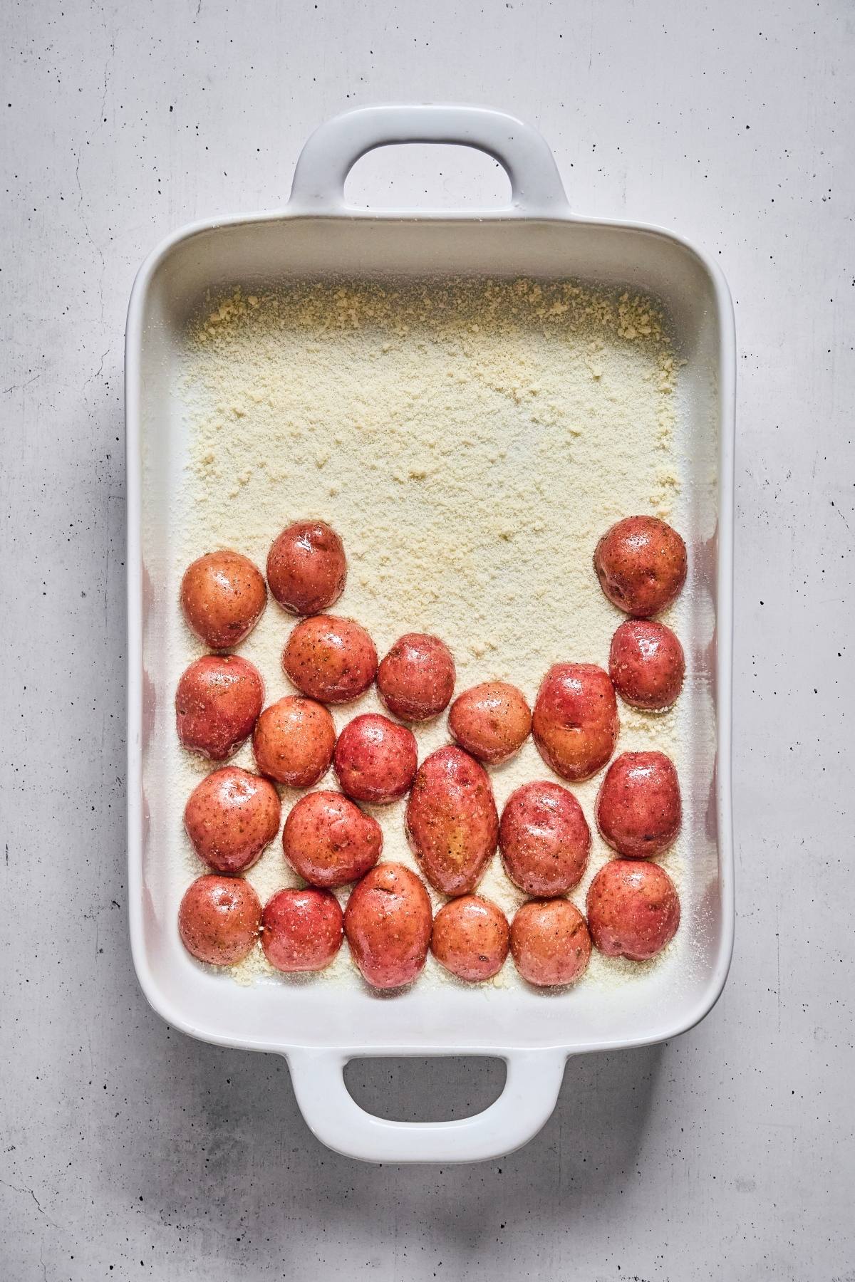 Red potatoes in a white baking dish on a layer of grated cheese or breadcrumbs, ready to be baked.