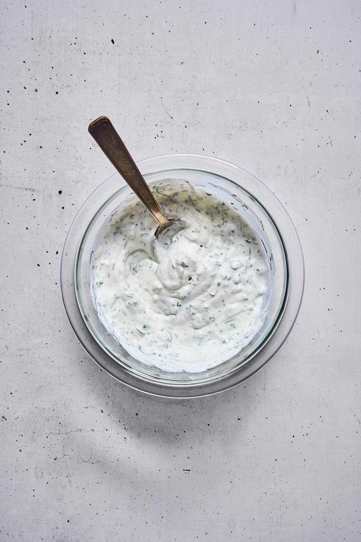 Glass bowl of creamy white sauce with herbs, stirred with a spoon, on a light textured surface.