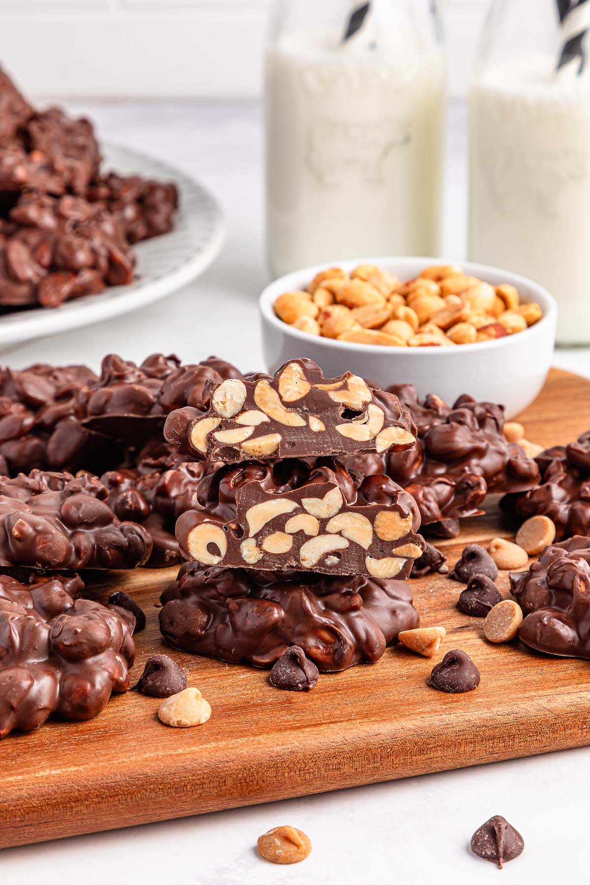 Crockpot Candy chocolate peanut clusters on a wooden board, with peanuts, chocolate chips, and bottles of milk in the background.