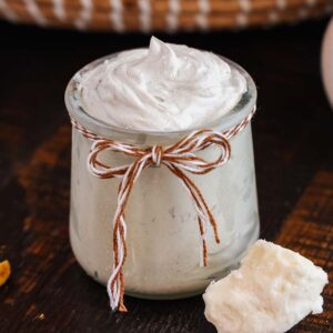 A small jar of homemade diaper rash cream, with a twine bow, placed on a wooden surface beside a chunk of shea butter.