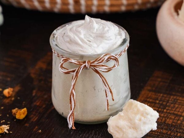 A small jar of homemade diaper rash cream, with a twine bow, placed on a wooden surface beside a chunk of shea butter.