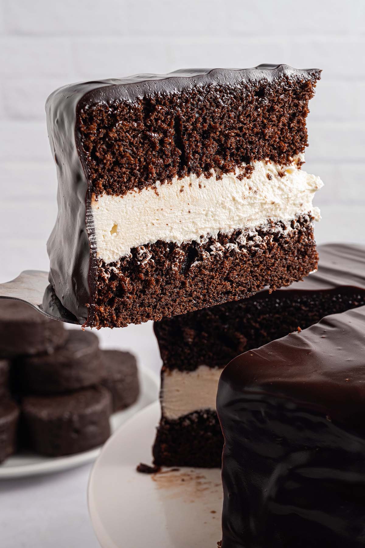 A slice of Ding Dong Cake with white cream filling and glossy chocolate frosting is being lifted from the cake.