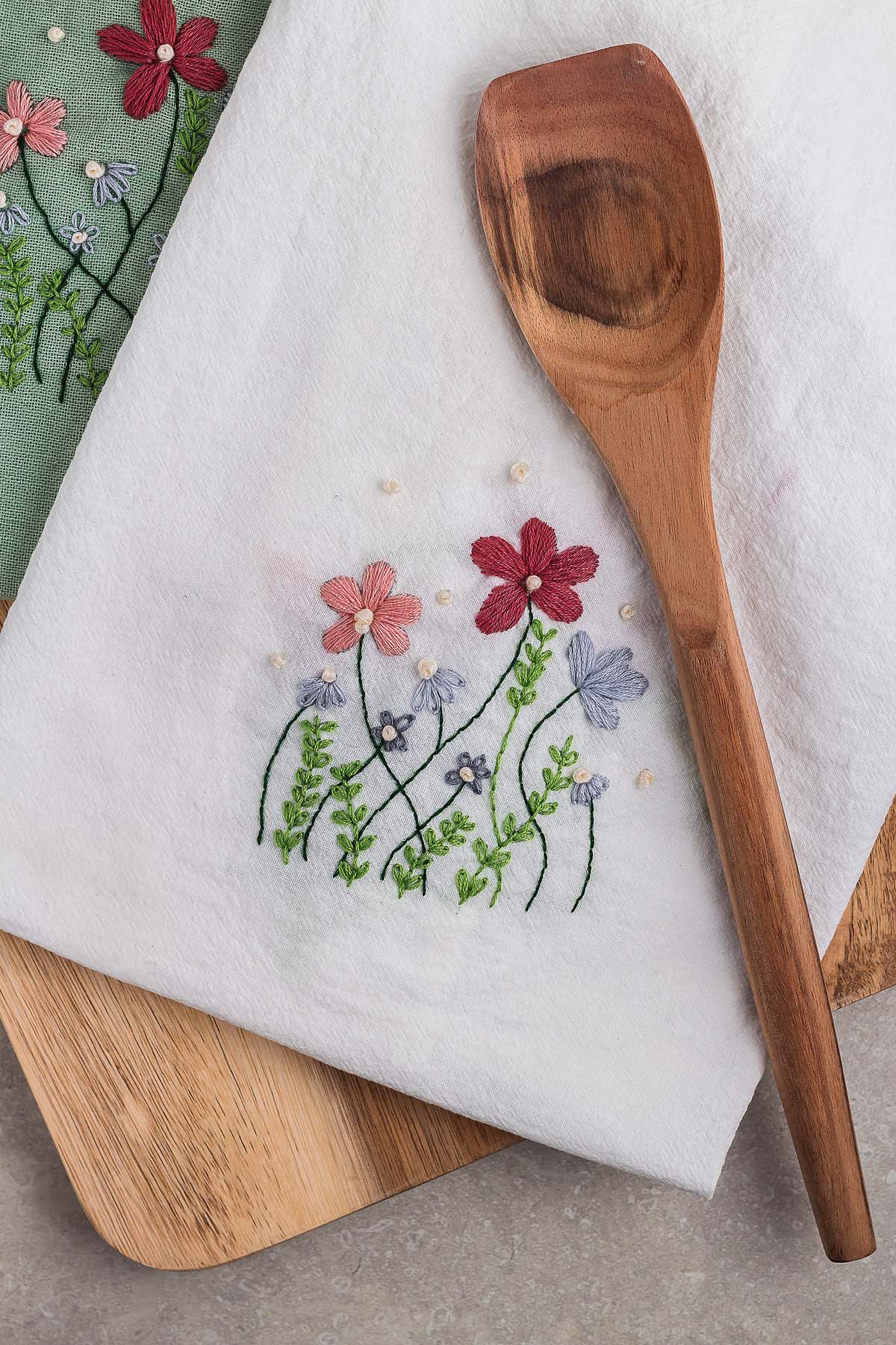Wooden spoon on a white cloth with colorful embroidered flowers, showcasing DIY embroidered flour sack towels, placed on wooden boards.