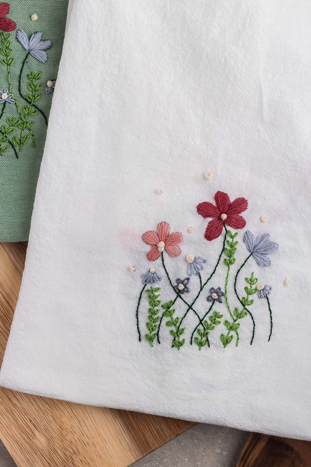 White fabric with colorful hand-embroidered flowers and green leaves, these DIY embroidered flour sack towels are beautifully displayed on a wooden surface.