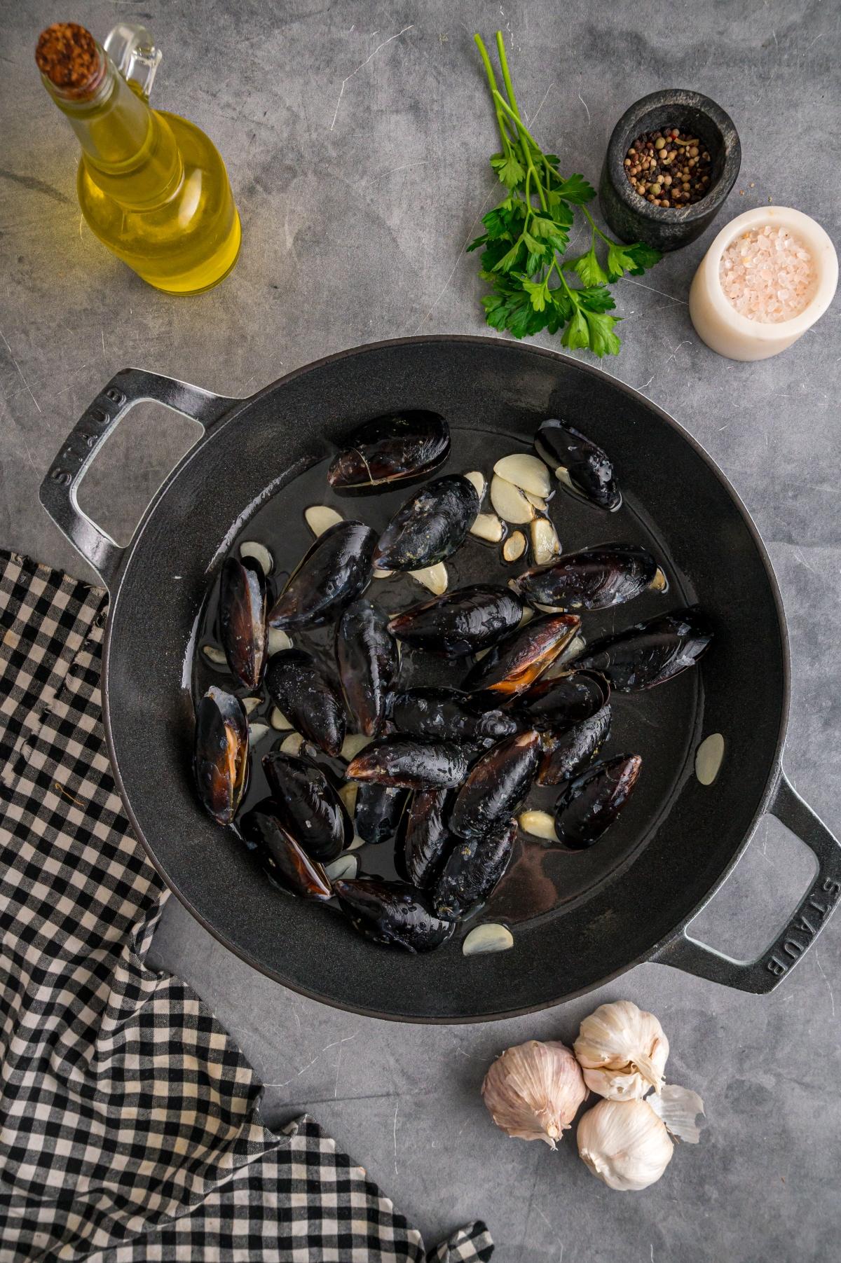 Black mussels with sliced garlic in a pan, surrounded by oil, parsley, seasonings, garlic, and a checkered cloth.