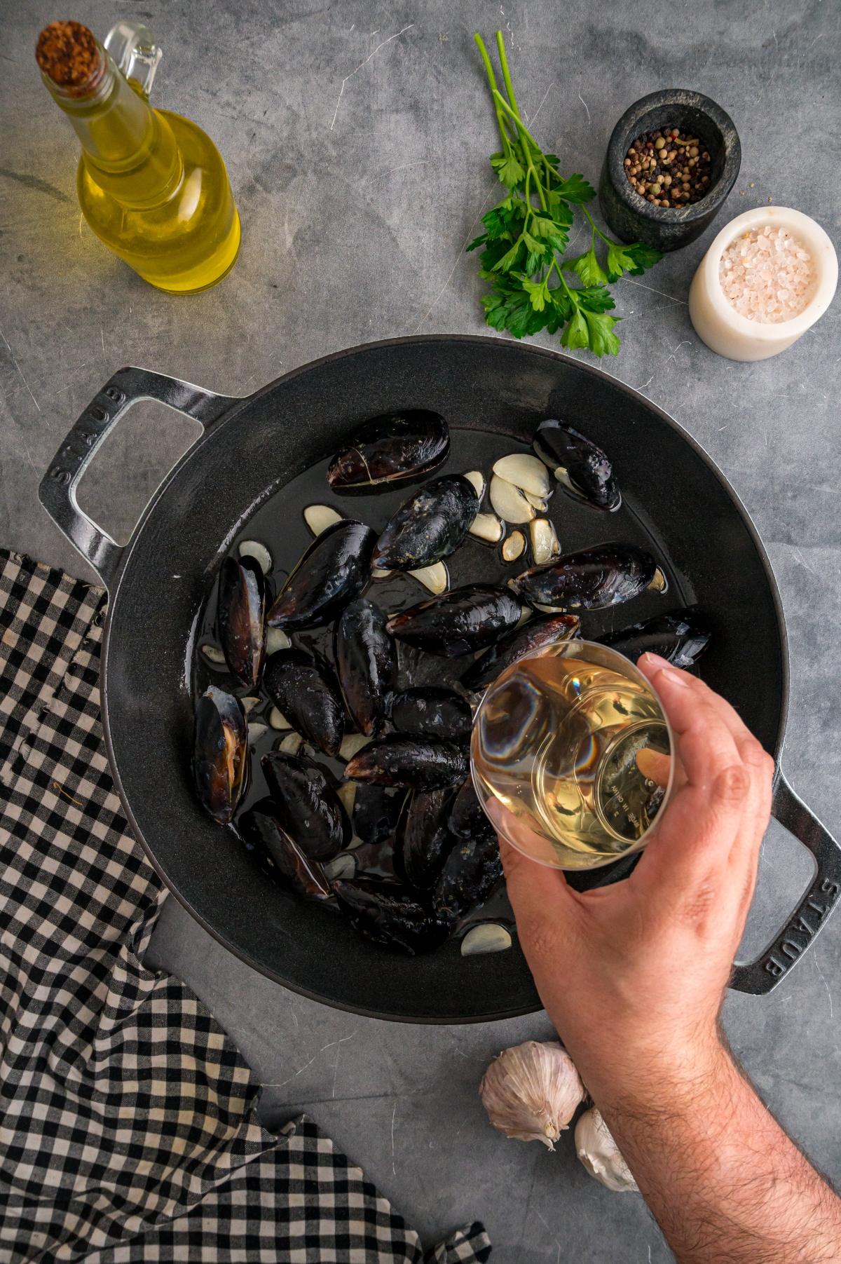A hand pours white wine over mussels and garlic in a black pan, surrounded by herbs and seasonings.
