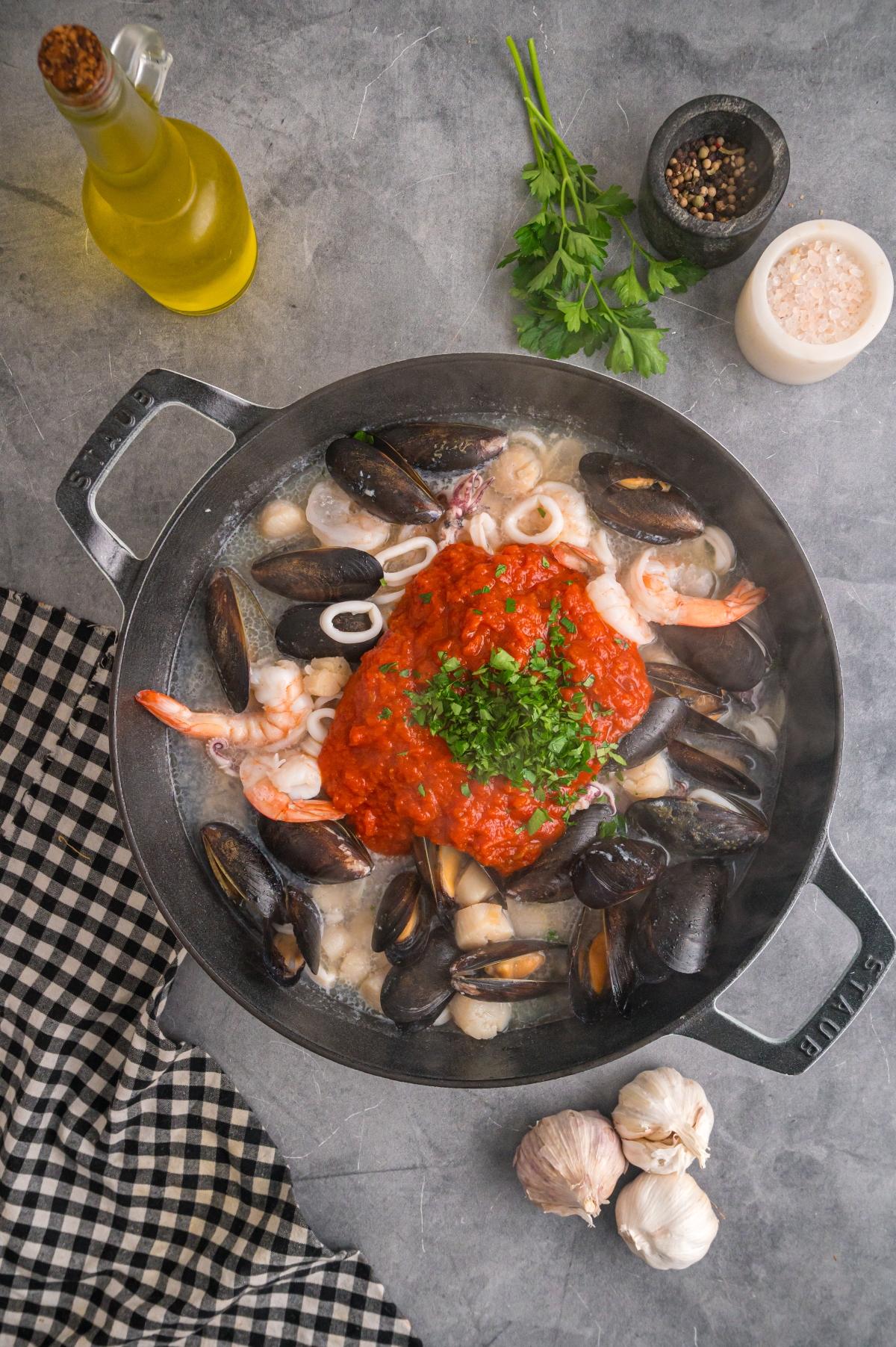 A skillet with assorted seafood, tomato sauce, and herbs, surrounded by seasonings and a checked cloth.