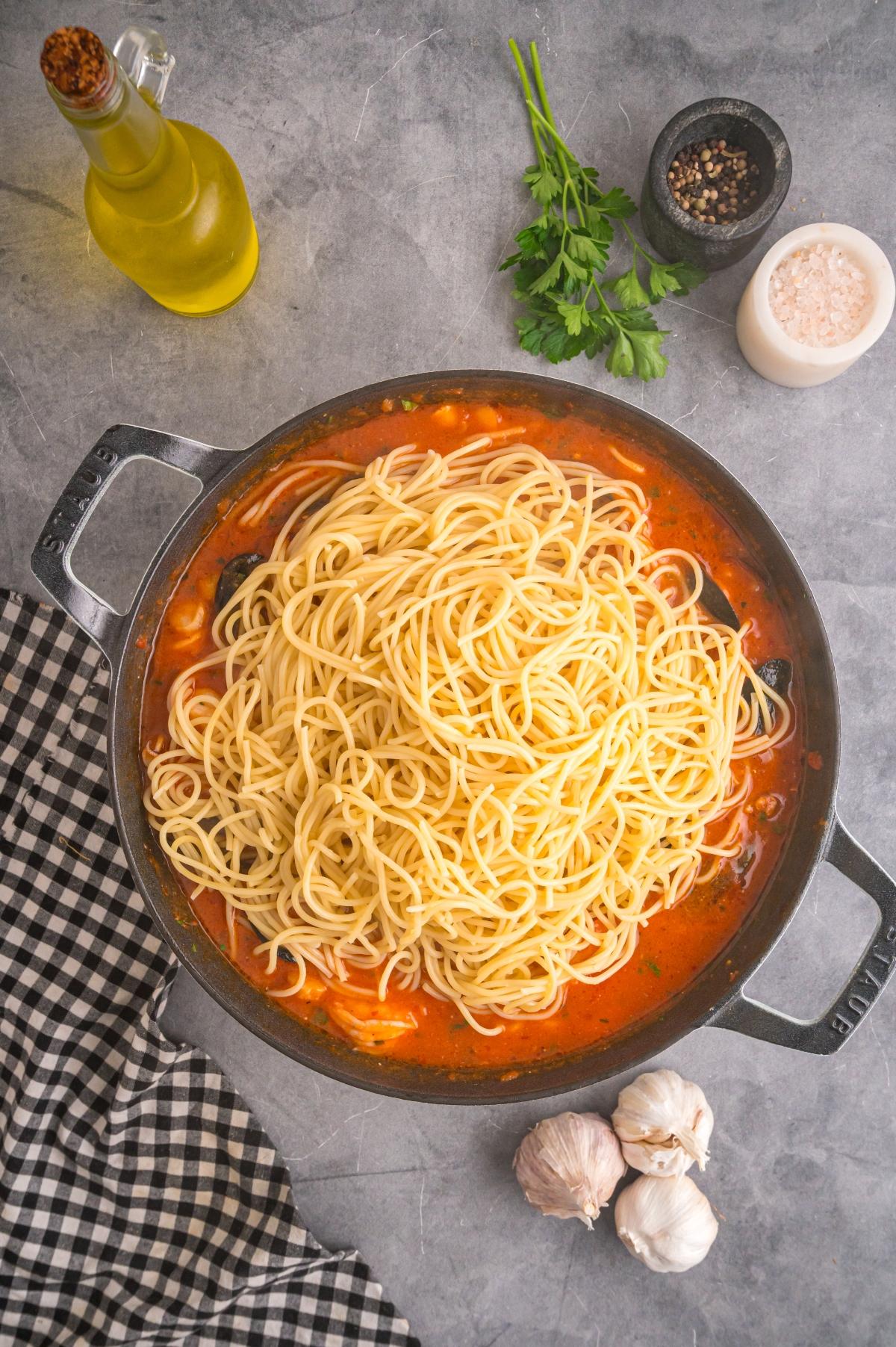A pan of cooked spaghetti on tomato sauce, surrounded by garlic, parsley, olive oil, salt, and pepper.
