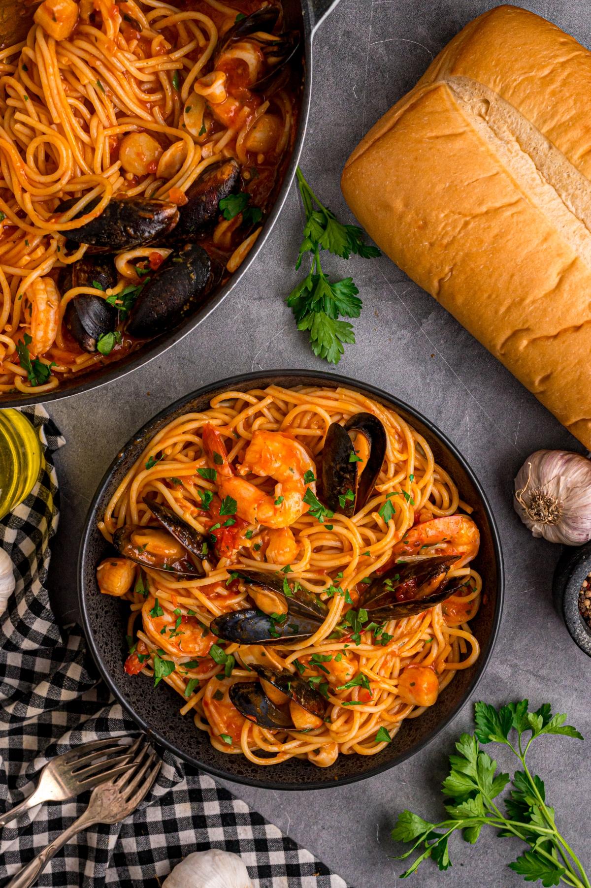 A bowl of seafood spaghetti with shrimp and mussels, next to bread, garlic, and fresh parsley on a table.