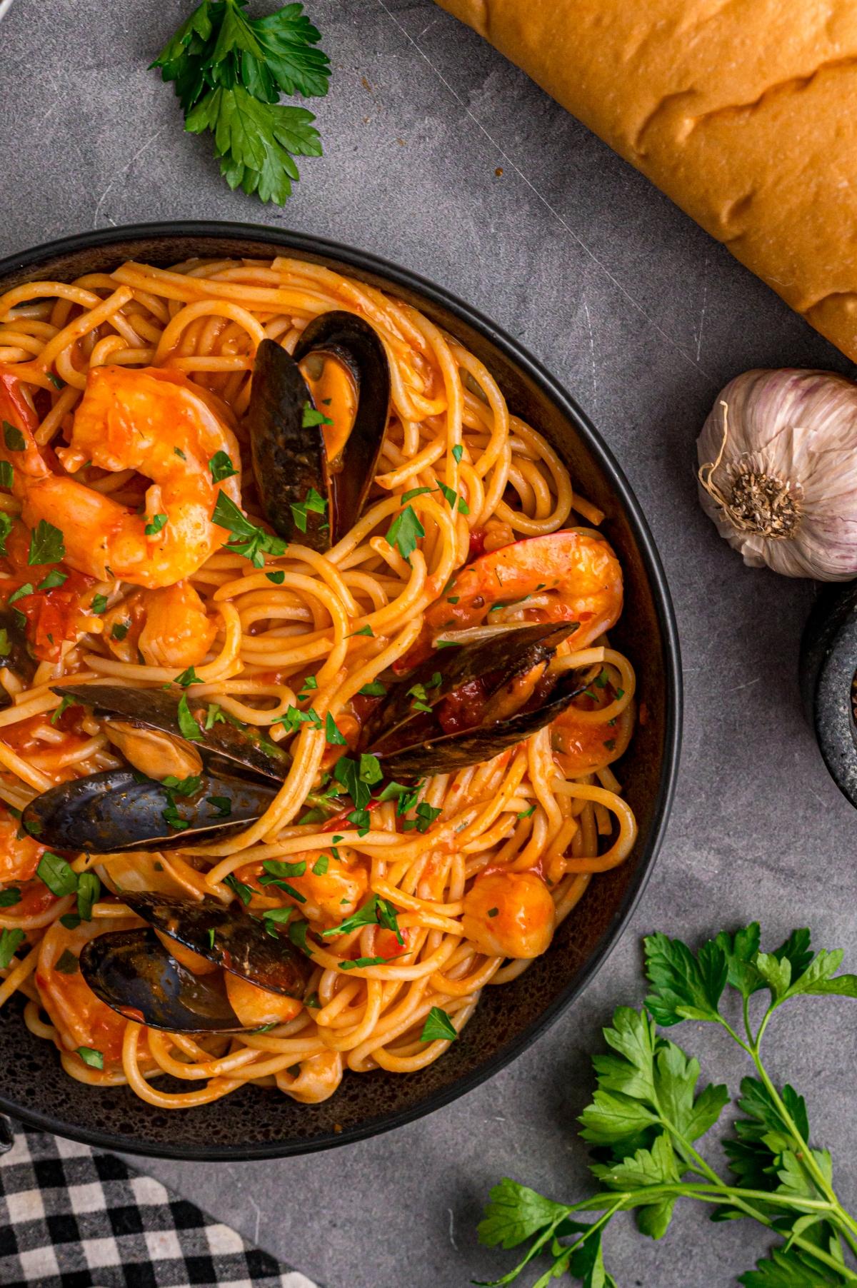 A bowl of seafood spaghetti with shrimp and mussels, garnished with parsley, next to bread and garlic.
