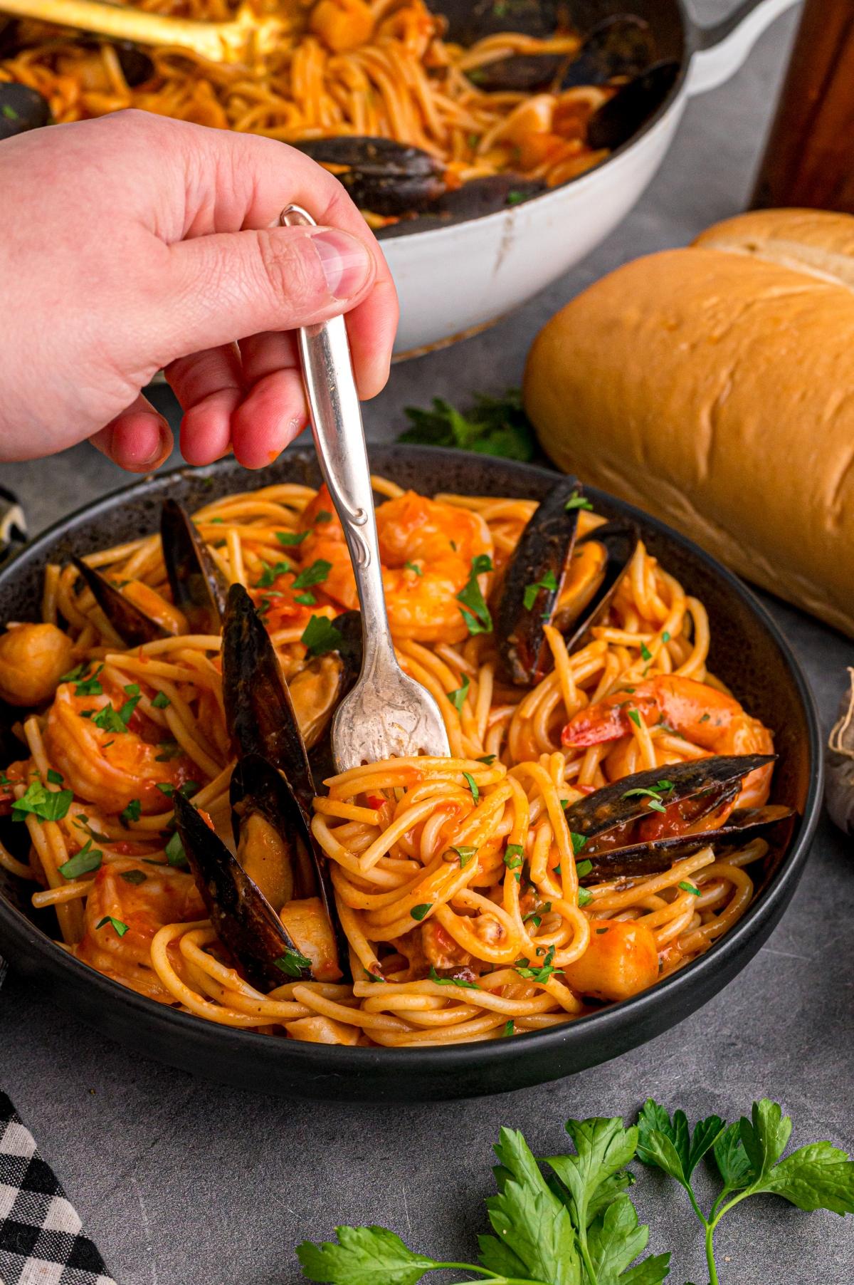 A hand twirls spaghetti with seafood in a black bowl, with bread and herbs nearby on the table.