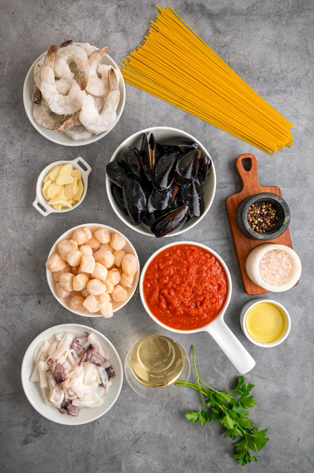 Various seafood, pasta, tomato sauce, seasonings, wine, oil, and parsley arranged on a gray surface.