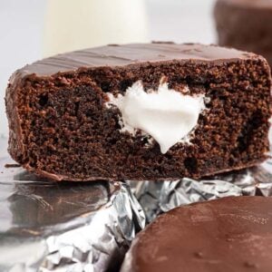Homemade Ding Dongs chocolate snack cake with cream filling, cut in half, rests on foil-wrapped cakes; more cakes in the background.