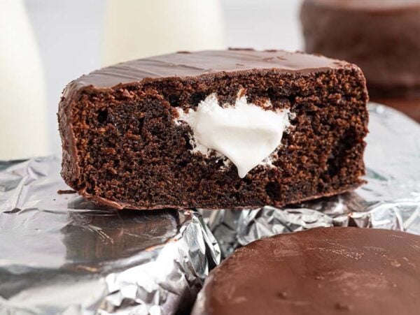 Homemade Ding Dongs chocolate snack cake with cream filling, cut in half, rests on foil-wrapped cakes; more cakes in the background.