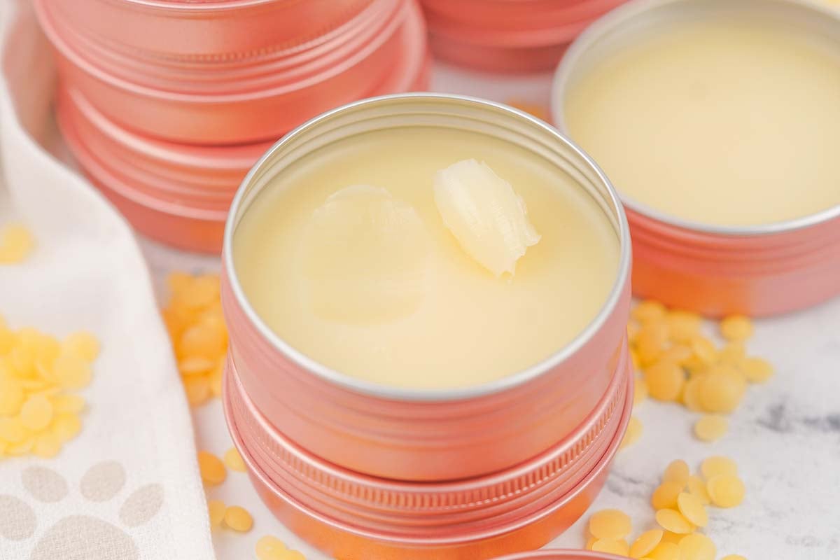 A small tin of creamy Dog Paw Balm with a swipe taken, surrounded by yellow pellets and other tins.