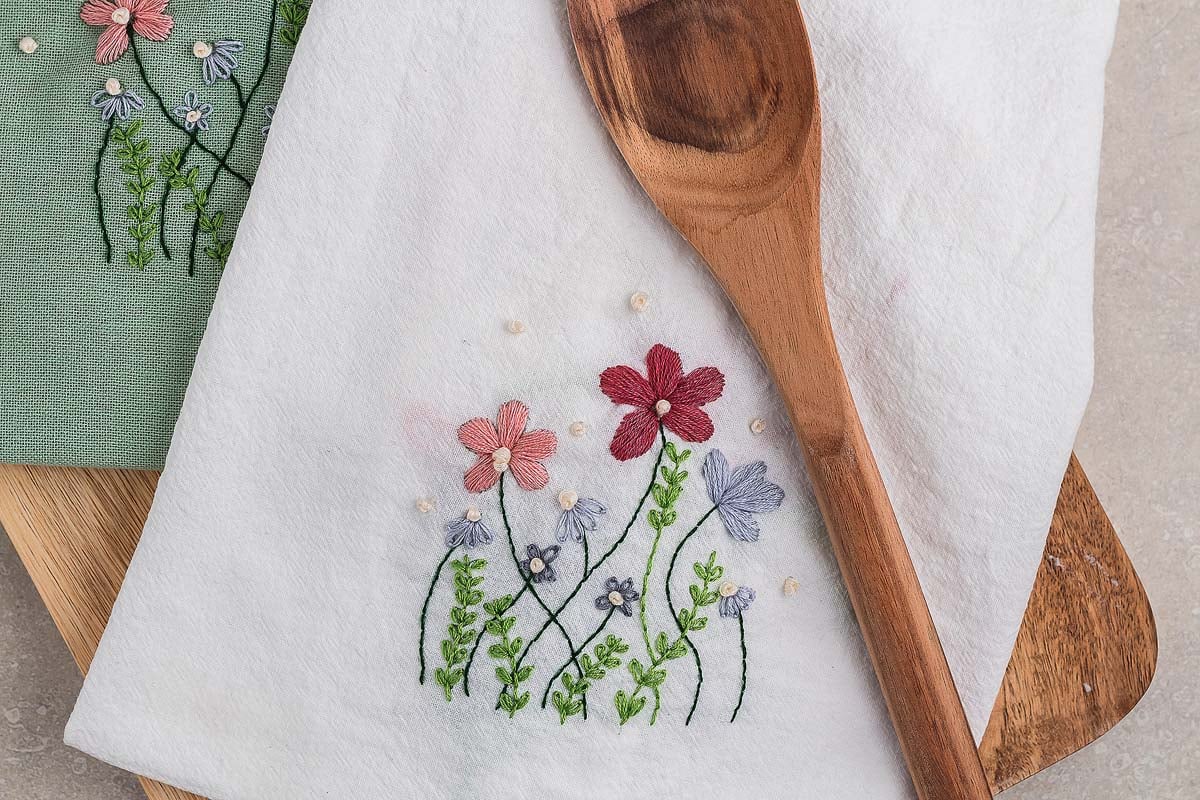 A wooden spoon rests on a DIY embroidered flour sack towel, its white cloth adorned with colorful flowers and green leaves.