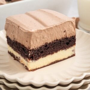 A slice of layered chocolate and vanilla Italian Love Cake with creamy topping sits on stacked plates, next to a glass of milk.