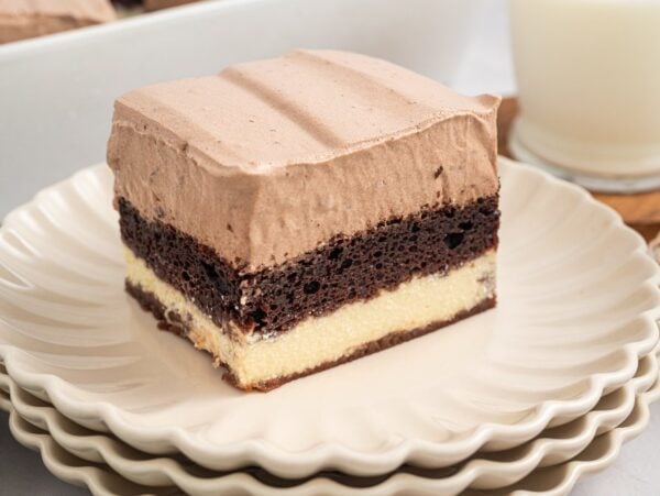 A slice of layered chocolate and vanilla Italian Love Cake with creamy topping sits on stacked plates, next to a glass of milk.