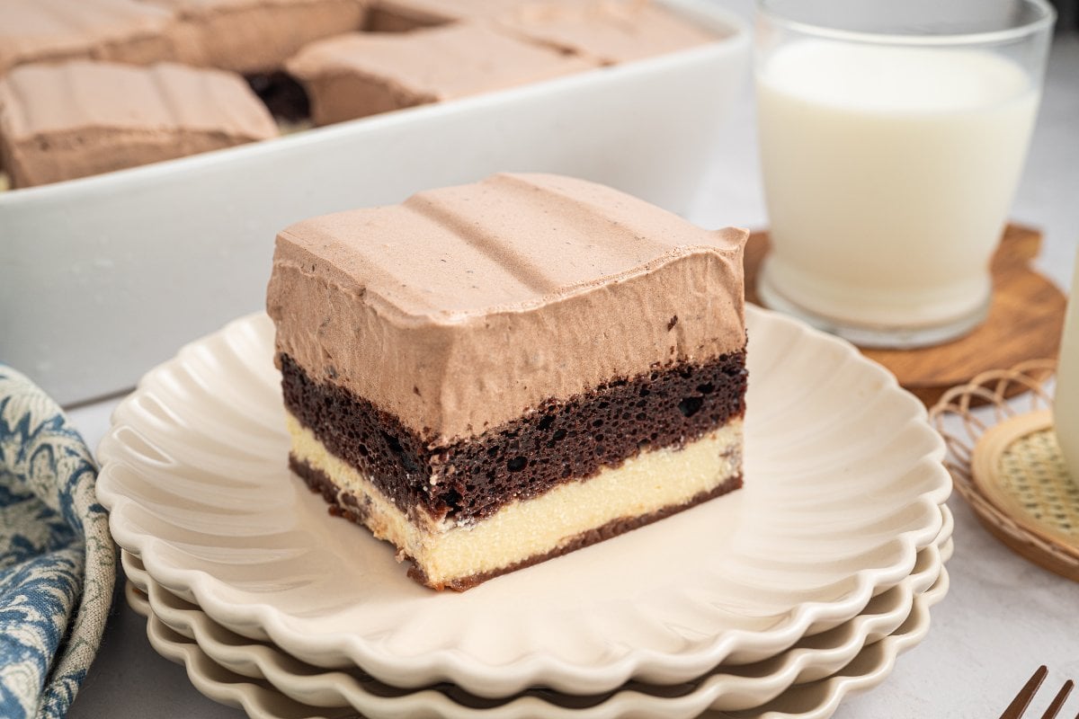 A slice of layered Italian Love Cake, featuring chocolate and vanilla with rich chocolate frosting, sits on a stack of plates next to a glass of milk.